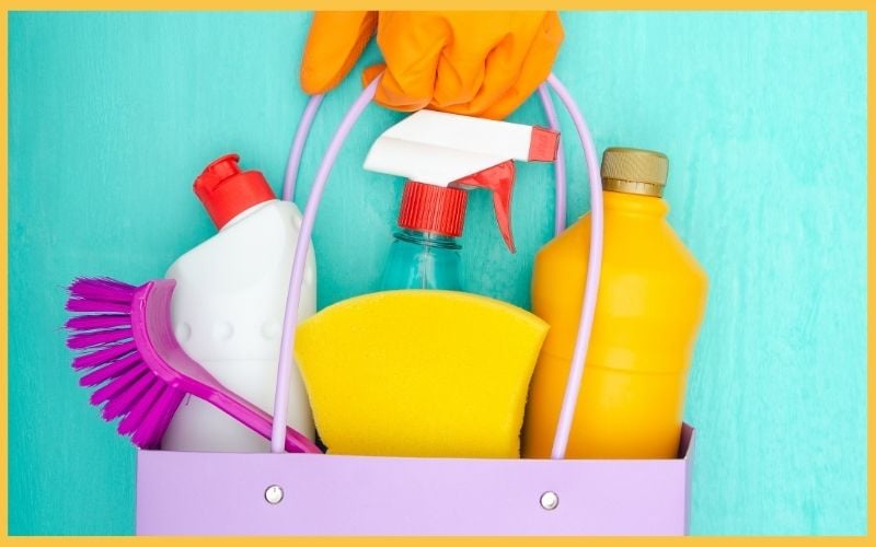A hand in an orange glove holds a spray bottle above a lavender cleaning caddy filled with various cleaning products and a sponge, to clean home like a pro.