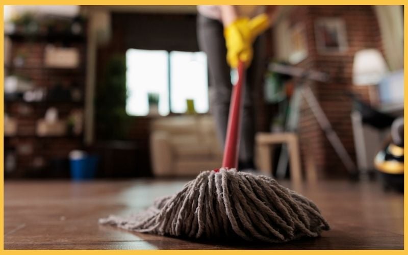 A person with yellow rubber gloves mops a wooden floor in a cozy living room, blurred background featuring furniture and plants.