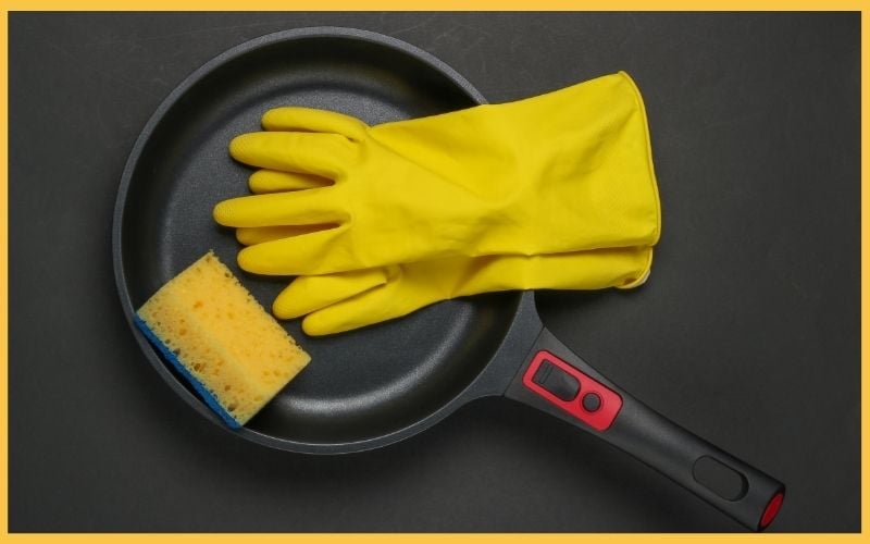 A black frying pan with a pair of yellow rubber gloves and a blue and yellow sponge resting on its surface, set against a dark background.