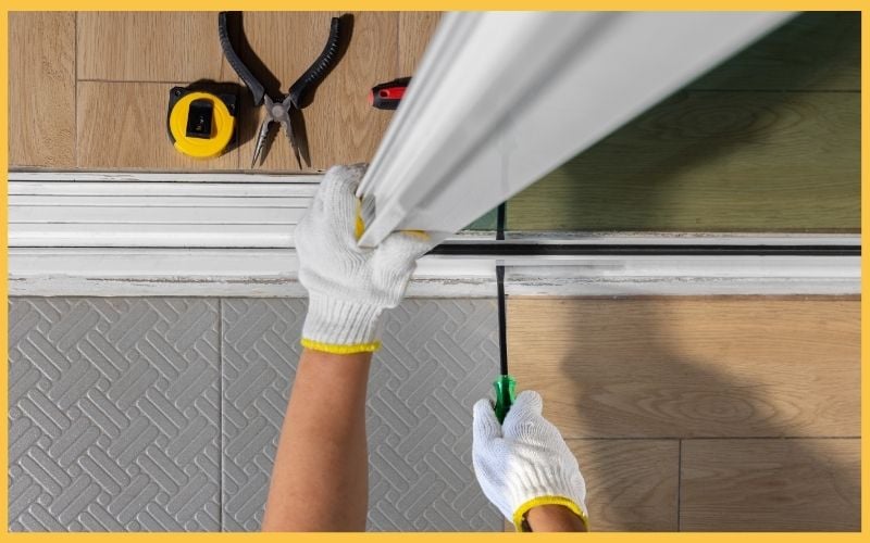 A person in white gloves uses a screwdriver to adjust a door frame, surrounded by tools like pliers and a tape measure on a wooden floor.