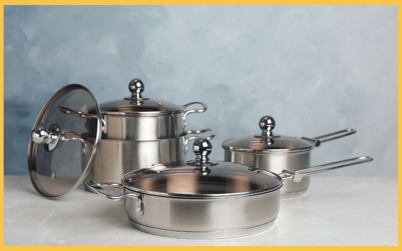 Three stainless steel cooking pots, one with a lid, arranged on a light surface against a soft blue backdrop.