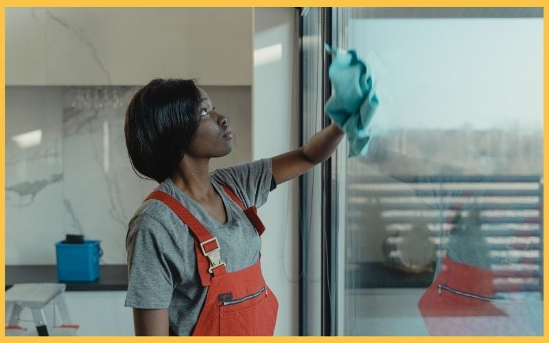 A person wearing a gray shirt and red overalls wipes a large glass door with a blue cloth, showcasing a bright living space.