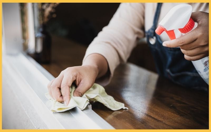 A person cleaning a wooden surface with a green cloth and a spray bottle, focusing on maintaining a tidy space.