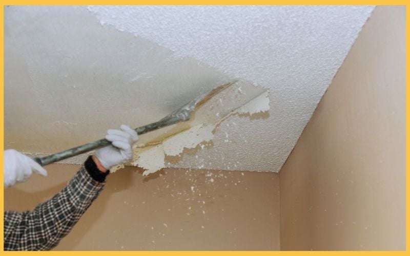 A person with gloved hands scraping popcorn texture from a ceiling in a beige room, debris falling into the air.