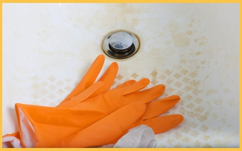 A pair of orange rubber gloves rests on a stained sink with a chrome drain, indicating a cleaning task underway.