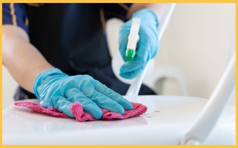 A person wearing blue gloves cleans a white chair with a pink cloth and spray bottle, focusing on a spot on the seat.