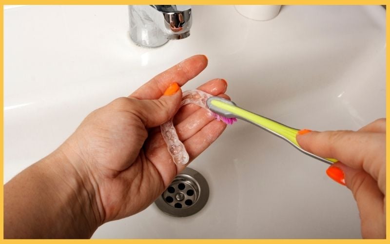 Person brushing a clear dental retainer with a toothbrush over a sink, demonstrating dental hygiene routine.