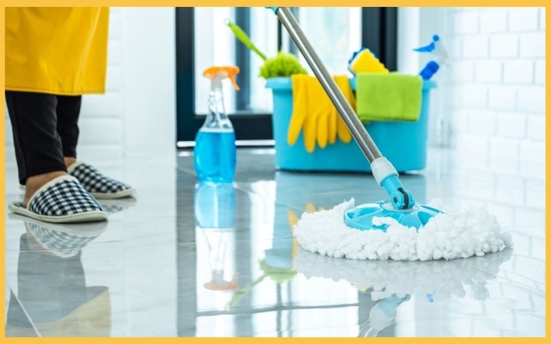 A person mops a shiny floor wearing checkered slippers, with cleaning supplies like a spray bottle and bucket in the background.