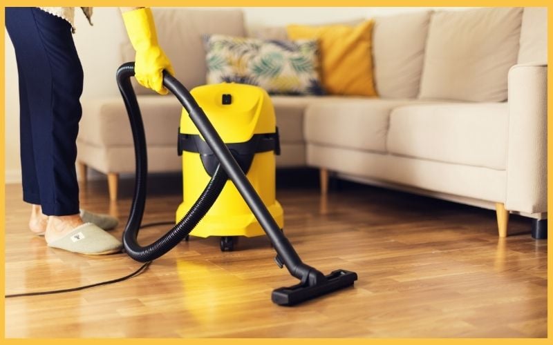 A person in yellow gloves vacuums a wooden floor near a beige sofa, using a bright yellow vacuum cleaner.