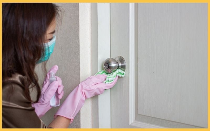 A woman in a mask and pink gloves cleans a door knob with a cloth while holding a spray bottle, emphasizing hygiene and care.