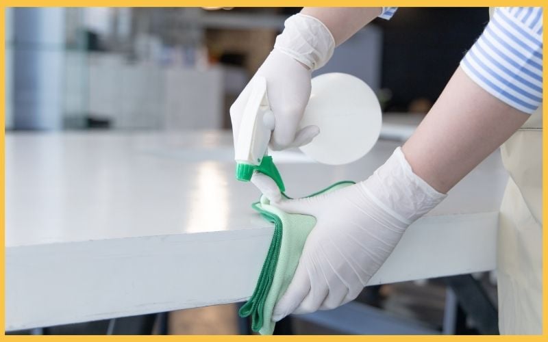 A person in gloves sprays a disinfectant on a table while wiping it clean with a green cloth in a bright, modern setting.