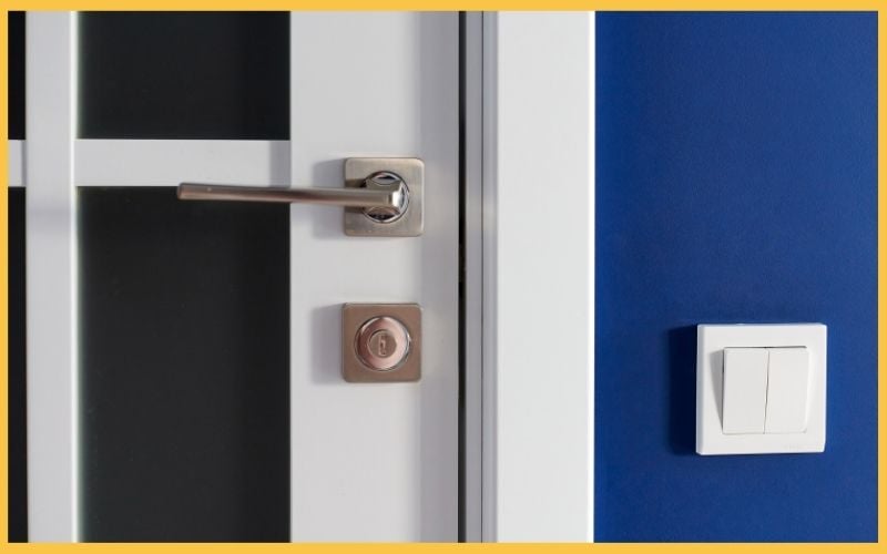 Close-up of a white door with a sleek silver handle and a cylinder lock, beside a light switch on a blue wall.