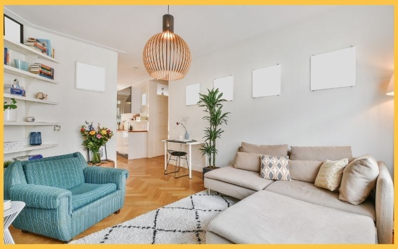 Cozy living room with a blue armchair, beige sofa, and modern decor, featuring a pendant light and a plant by a bright kitchen doorway.