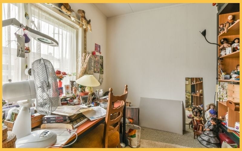 A cluttered room with a desk, fan, lamp, books, and shelves filled with dolls, reflecting a cozy, eclectic atmosphere.