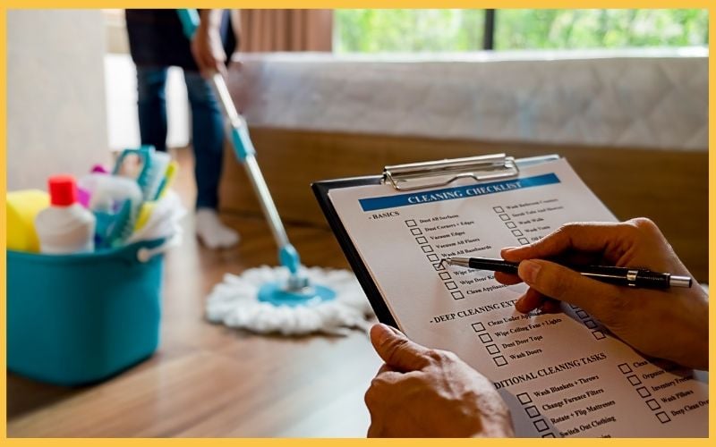 A person holds a cleaning checklist while another mops the floor, with a bucket of cleaning supplies nearby.