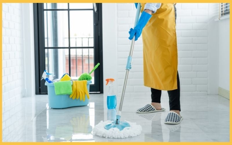  A person in a yellow apron and blue gloves mops a shiny floor beside a bucket filled with cleaning supplies.