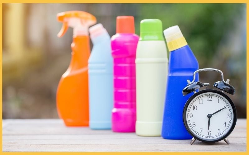 A lineup of colorful cleaning bottles next to a black alarm clock on a wooden surface, with a blurred background.