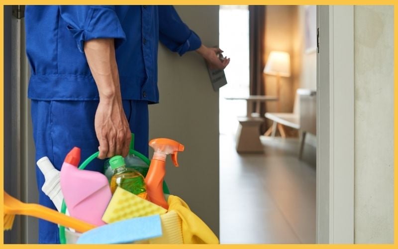 A person in a blue uniform holds a cleaning supplies basket, opening a door to a well-lit interior space.