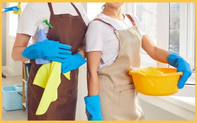 Two women in latex gloves and aprons hold cleaning supplies. One has a spray bottle and cloth, while the other holds a yellow bowl.