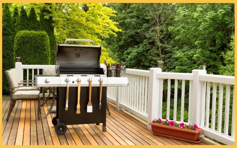 A grill on a wooden deck with cooking utensils neatly hanging, surrounded by lush greenery and a planter of flowers.