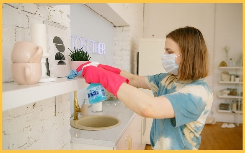 A person in colorful attire cleans a shelf with a spray bottle and cloth, surrounded by decorative items in a bright, modern kitchen.