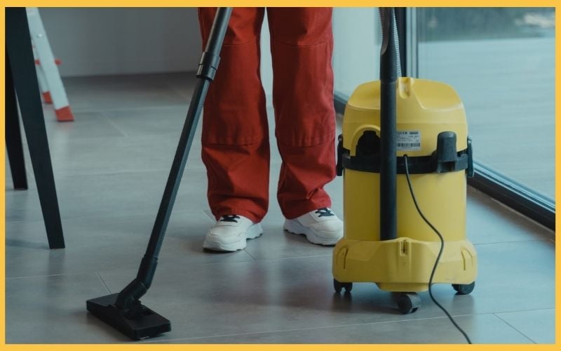 A person in red pants cleans the floor with a vacuum, next to a yellow vacuum cleaner, beside a large window.