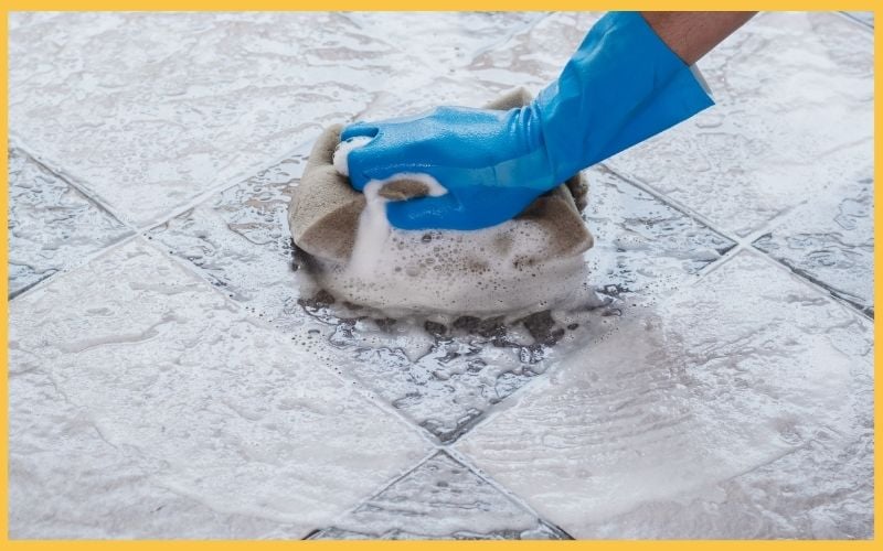 A gloved hand scrubs a textured tile floor with a soap-filled sponge, creating bubbles amidst a sparkling clean surface.