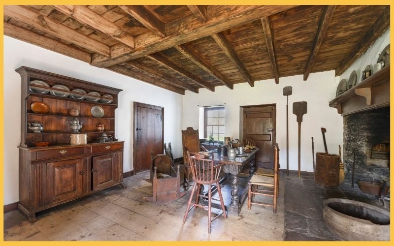 A rustic kitchen with wooden beams, a large dining table, antique chairs, and a sideboard displaying vintage cookware and utensils.