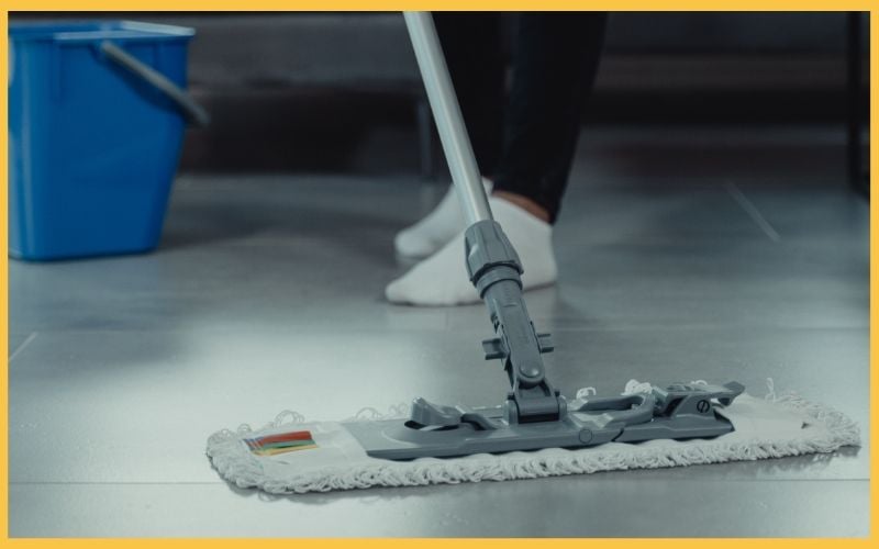 A person mops a floor with a microfiber mop, standing beside a blue bucket, wearing socks on shiny tile flooring.