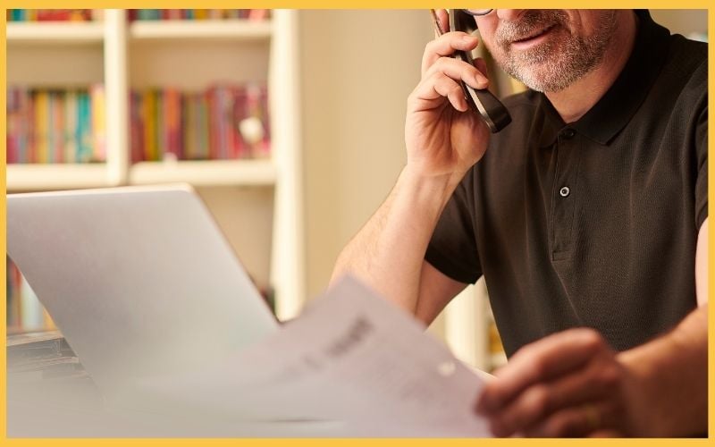 A man in a black polo shirt holds a phone to his ear while reviewing documents beside a laptop, with colorful books in the background.