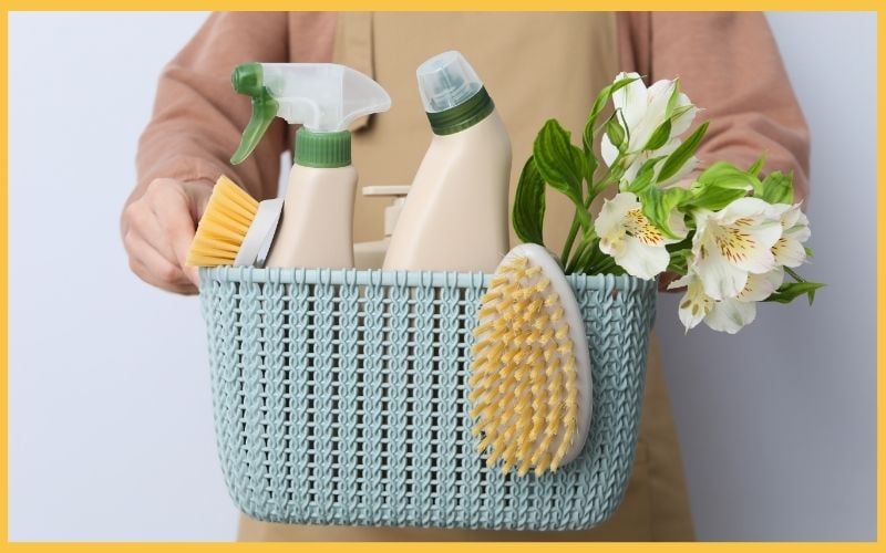 A person holds a teal basket filled with cleaning supplies, including spray bottles, a scrub brush, and a bouquet of flowers.