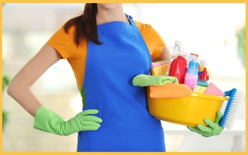 A woman in a blue apron and green gloves holds a yellow cleaning bucket filled with various cleaning supplies and colorful towels.