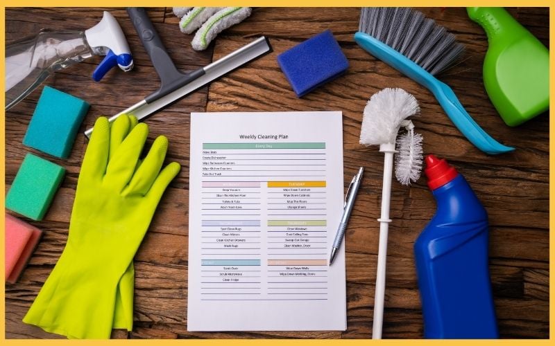 A weekly cleaning plan sheet surrounded by cleaning supplies: gloves, sponges, spray bottles, a brush, and a pen on a wooden surface.
