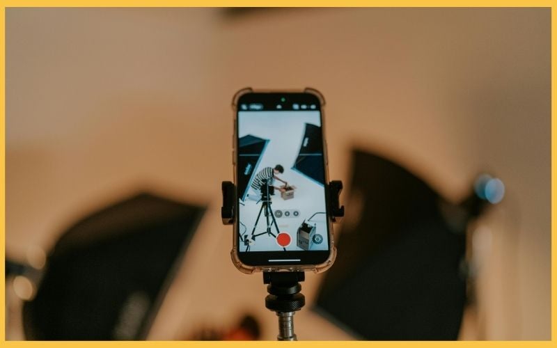 A smartphone mounted on a stand displays a recording scene of a person setting up a photoshoot with studio lights.